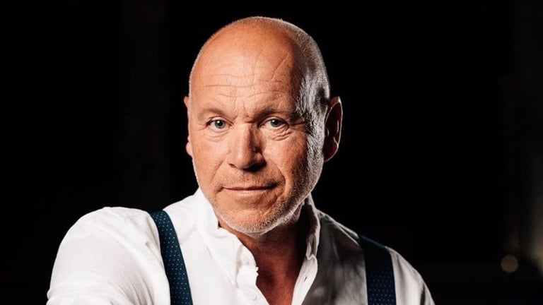 Professional headshot of a bald man wearing a white shirt and dark vest against a black background