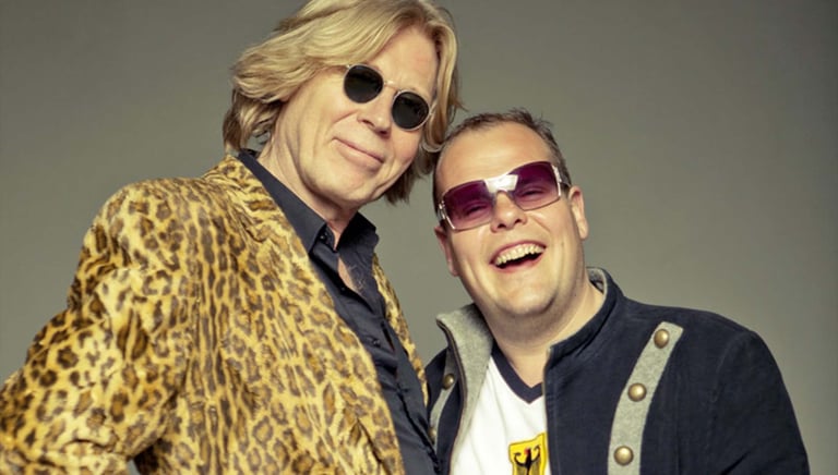 Two men wearing sunglasses pose together against a gray background. The man on the left wears a leopard print jacket and round sunglasses, while the man on the right wears a dark jacket and pink-tinted sunglasses.